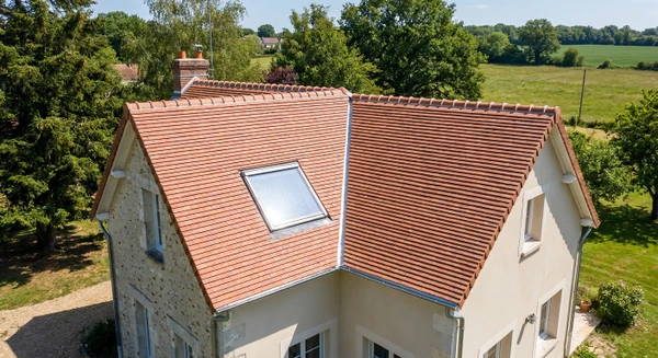 Photographie a&eacute;rienne ou vue en l&eacute;g&egrave;re plong&eacute;e d'une maison individuelle avec toit en tuiles ou ardoises dans un contexte rural ou p&eacute;ri-urbain typique des Yvelines, montrant clairement la toiture en bon &eacute;tat ou en cours de r&eacute;novation. L'image doit capturer l'essence du m&eacute;tier de couvreur-zingueur en mettant l'accent sur les &eacute;l&eacute;ments de toiture (couverture, goutti&egrave;res visibles, solins). Journ&eacute;e ensoleill&eacute;e, ciel d&eacute;gag&eacute;. Pas de personnes visibles. Composition professionnelle et nette.