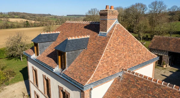 Photographie en vue a&eacute;rienne ou l&eacute;g&egrave;re plong&eacute;e d'une maison individuelle traditionnelle aux toits en tuiles rouges ou ardoises grises, situ&eacute;e dans un environnement rural ou p&eacute;ri-urbain des Yvelines. La maison doit &ecirc;tre clairement visible avec ses &eacute;l&eacute;ments de toiture en d&eacute;tail&nbsp;: goutti&egrave;res m&eacute;talliques, solins, ch&eacute;neaux bien visibles. Contexte paysager typique du Nord de l'&Icirc;le-de-France avec v&eacute;g&eacute;tation environnante. Jour ensoleill&eacute;, ciel d&eacute;gag&eacute;. Aucune personne visible. Composition nette et professionnelle mettant l'accent sur la structure du toit et les &eacute;l&eacute;ments de zinguerie.