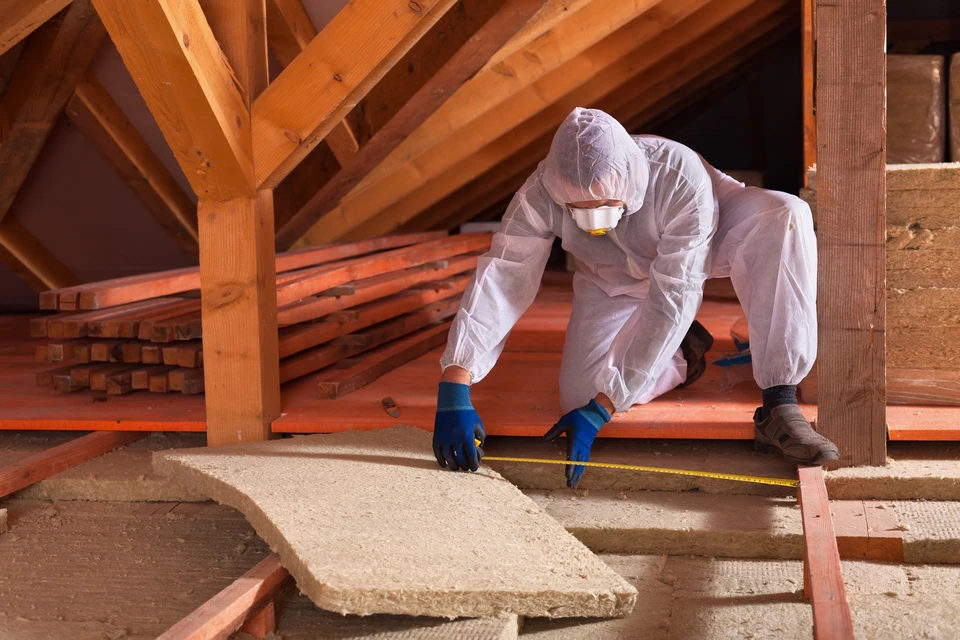 Professionnel en combinaison de protection blanche et gants bleus travaillant dans des combles, mesurant l'isolation avec un mètre ruban