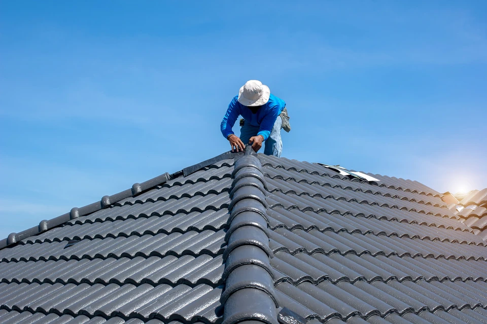 Ouvrier couvreur en tenue de protection travaillant sur toiture en tuiles mécaniques grises, ciel bleu dégagé
